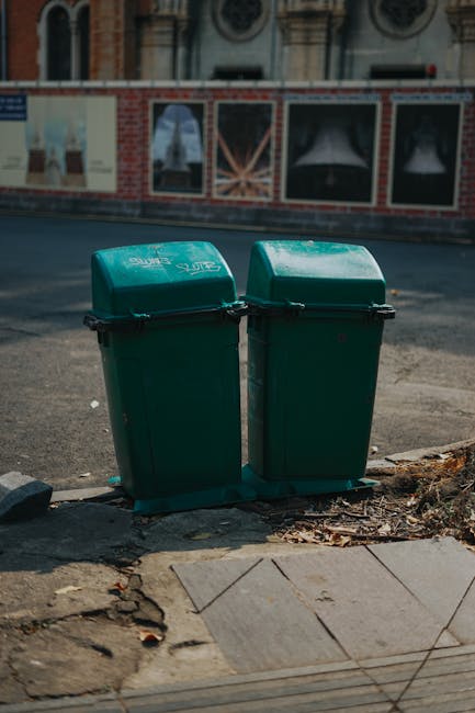 Two green outdoor waste bins with black latches positioned on a concrete sidewalk near the curb, adjacent to a patch of pavement with scattered leaves and debris. The bins are situated in front of a b